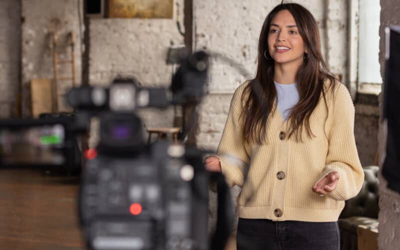 Brunette Woman in a Cream Cardigan stood in front of a filming camera