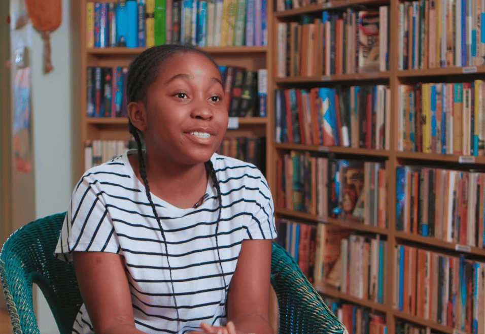 Young girl with braids sitting in a library speaking to someone off camera