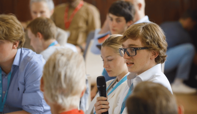 Young man in glasses holding a microphone and talking to a group of people