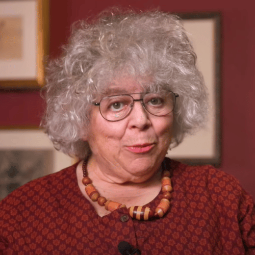 Miriam Margolyes in a red dress and wooden bead necklace, speaking to the camera
