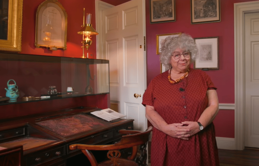 Miriam Margolyes stood in a drawing room wearing a red dress and looking at the camera