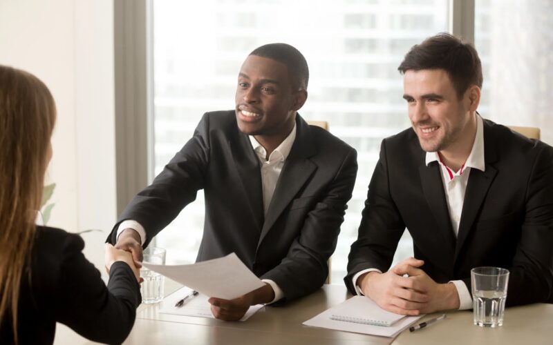 Two men in suits shake hands of woman across a table