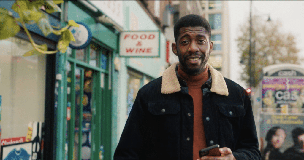 Presenter walks past food and wine shop holding phone.