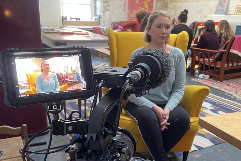 A mother, on a yellow chair, is being filmed and her image is shown in the camera monitor